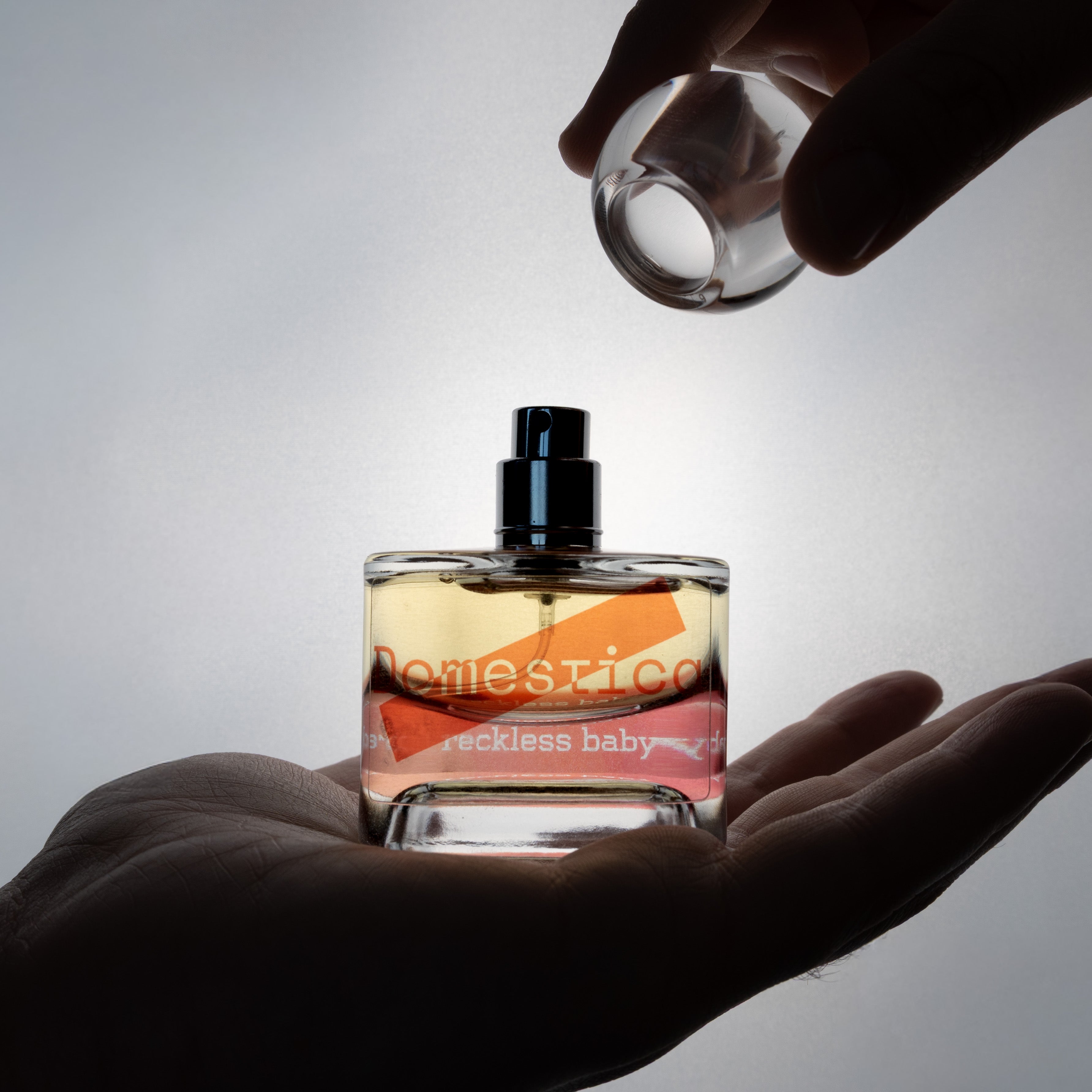 Perfume bottle labeled 'Domestica' and 'Reckless baby' held in a hand with a blurred background