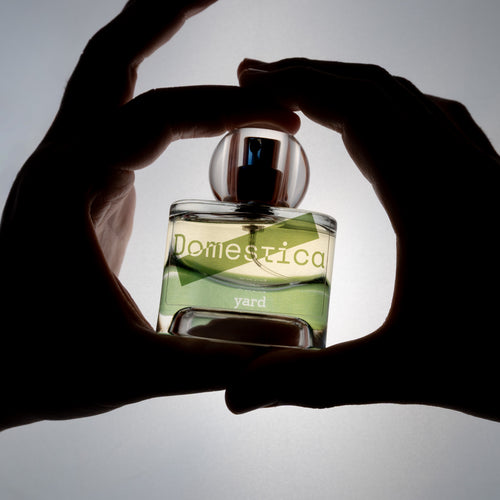 Two hands holding a glass bottle labeled 'Domestica' and 'Yard' against a gray background.