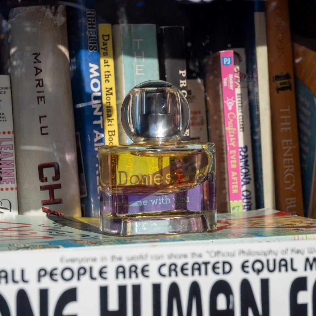 Perfume bottle labeled 'Domestica' and 'Home with you' on a bookshelf with books in the background.