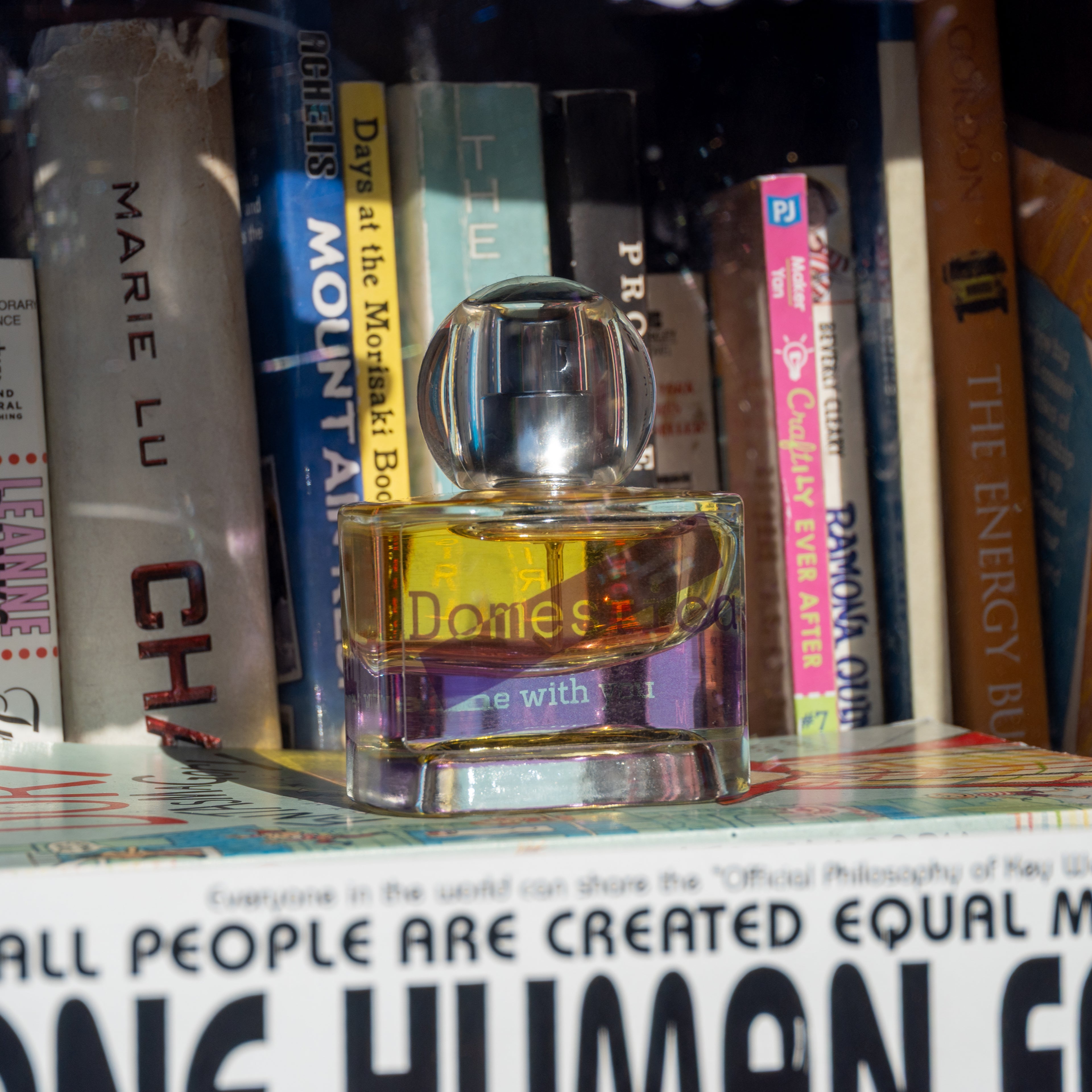 Perfume bottle labeled 'Domestica' and 'Home with you' on a bookshelf with books in the background.