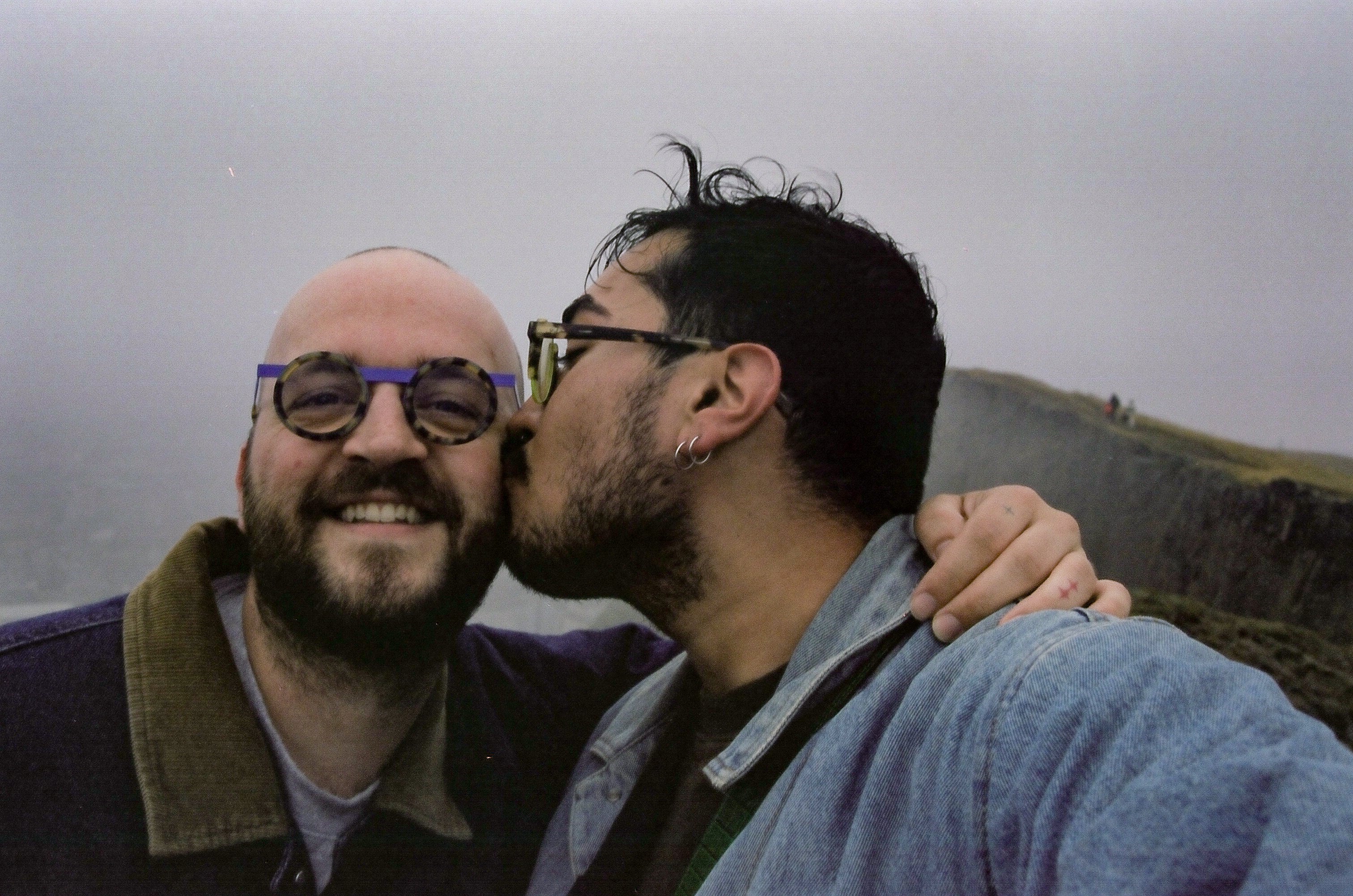 A man kissing another man on the cheek with a misty landscape in the background.